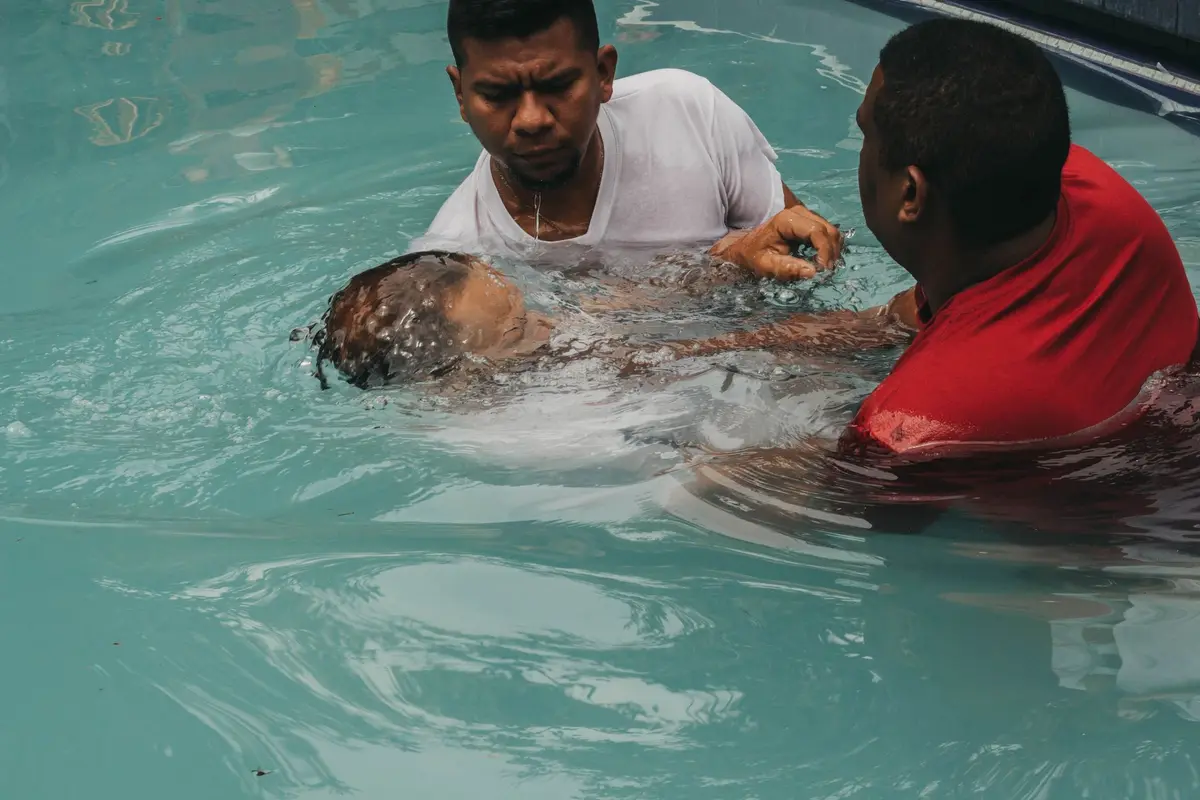 A pastor baptizing a young person in a pool while another man assists