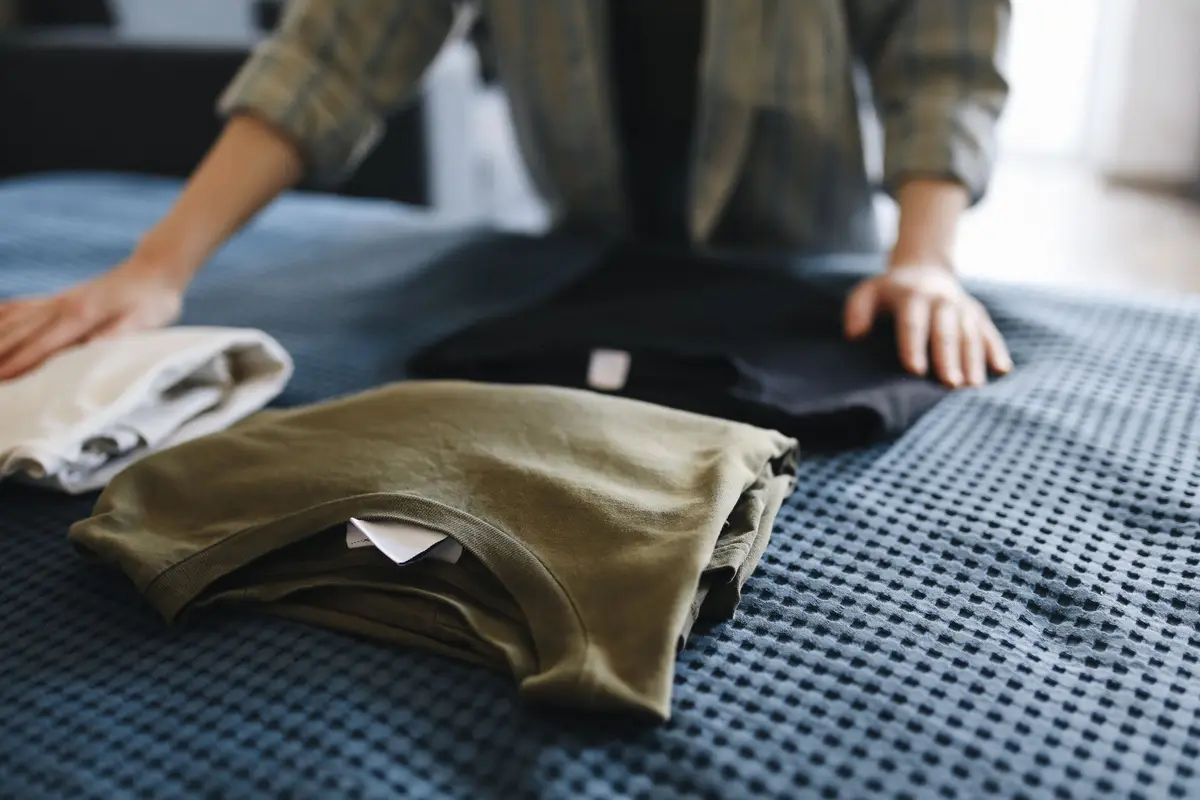 Hands folding a stack of t-shirts in olive, black, and white on a bed