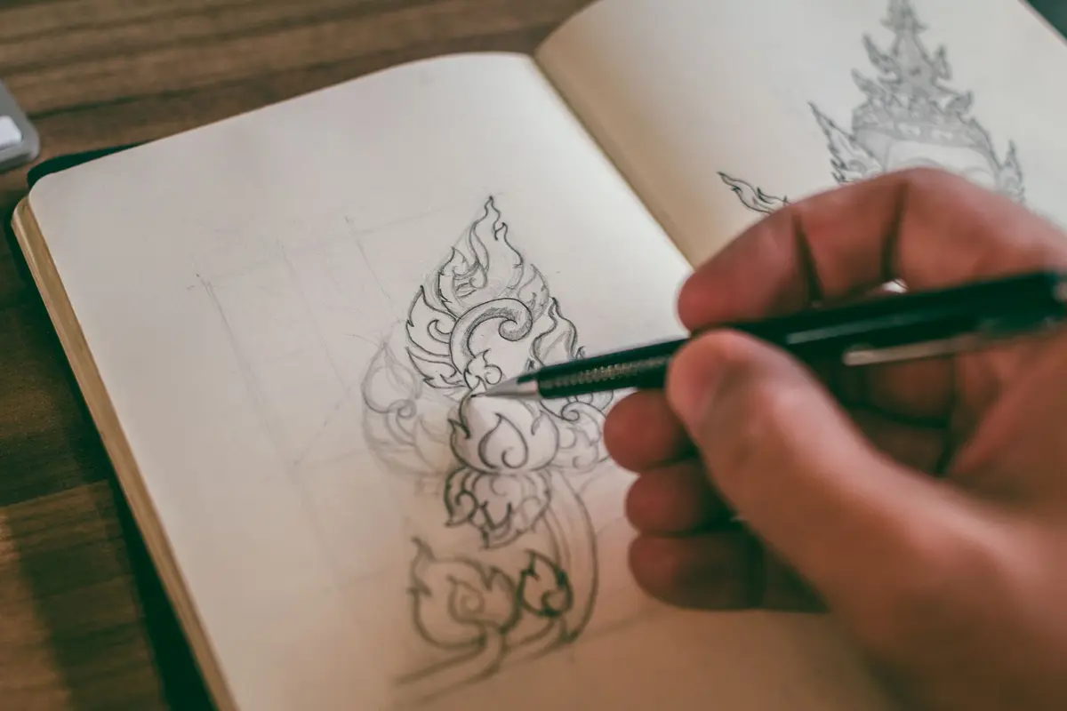 A hand sketching a detailed design in a sketchbook with a pen