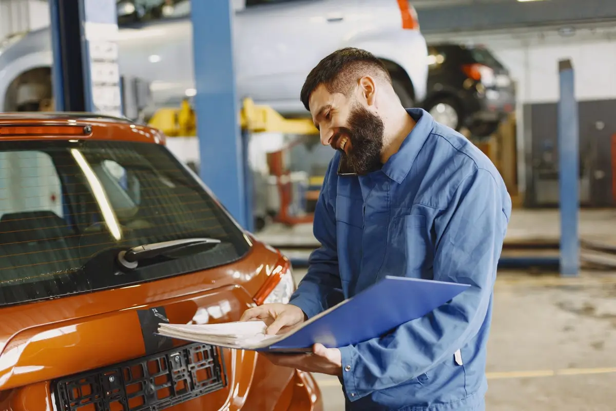 A mechanic in a blue work shirt checking a repair order on a clipboard beside a car