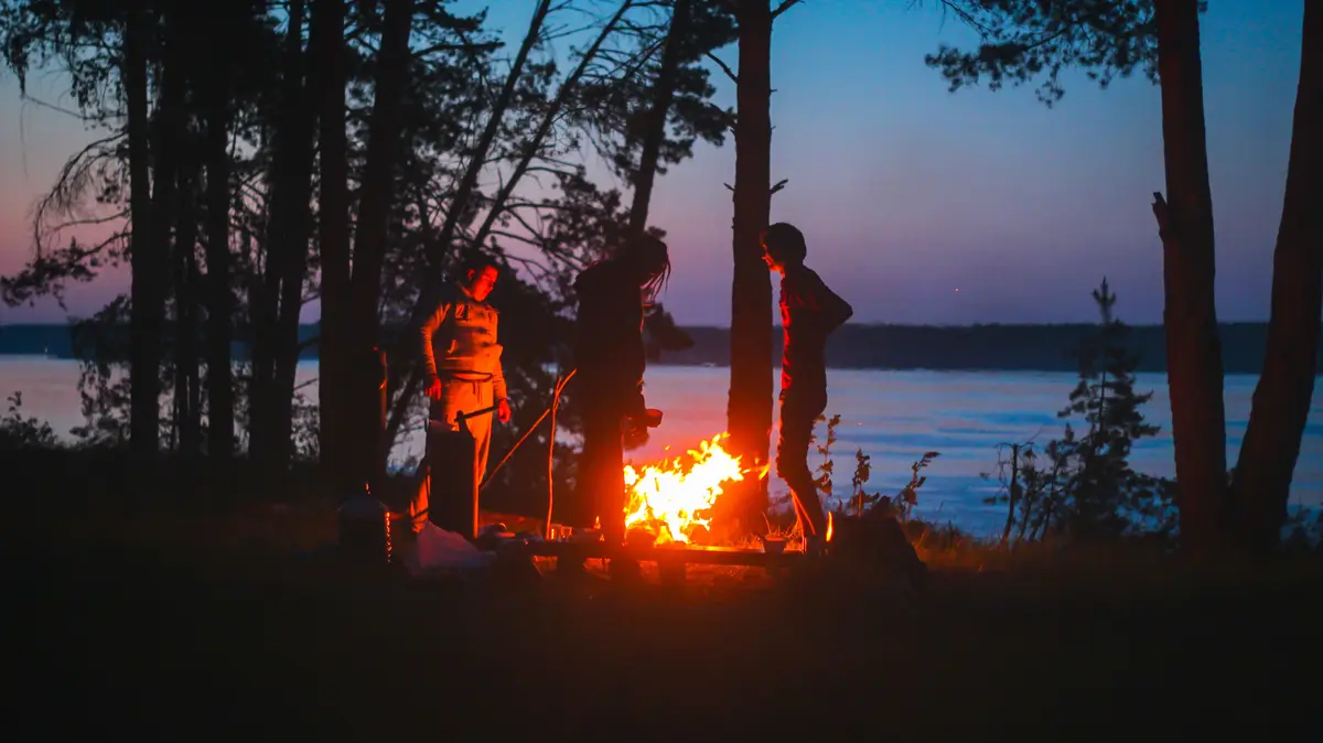 Silhouettes of campers standing around a campfire by a lake at dusk